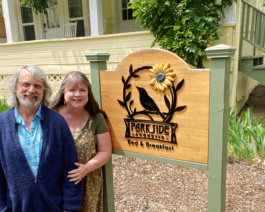 Parkside Bed & Breakfast sign in front of a yellow house, with owners posed on the left.